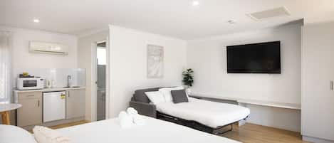 A modern hotel room with a bed, a small kitchenette, a flat-screen TV, and a potted plant.