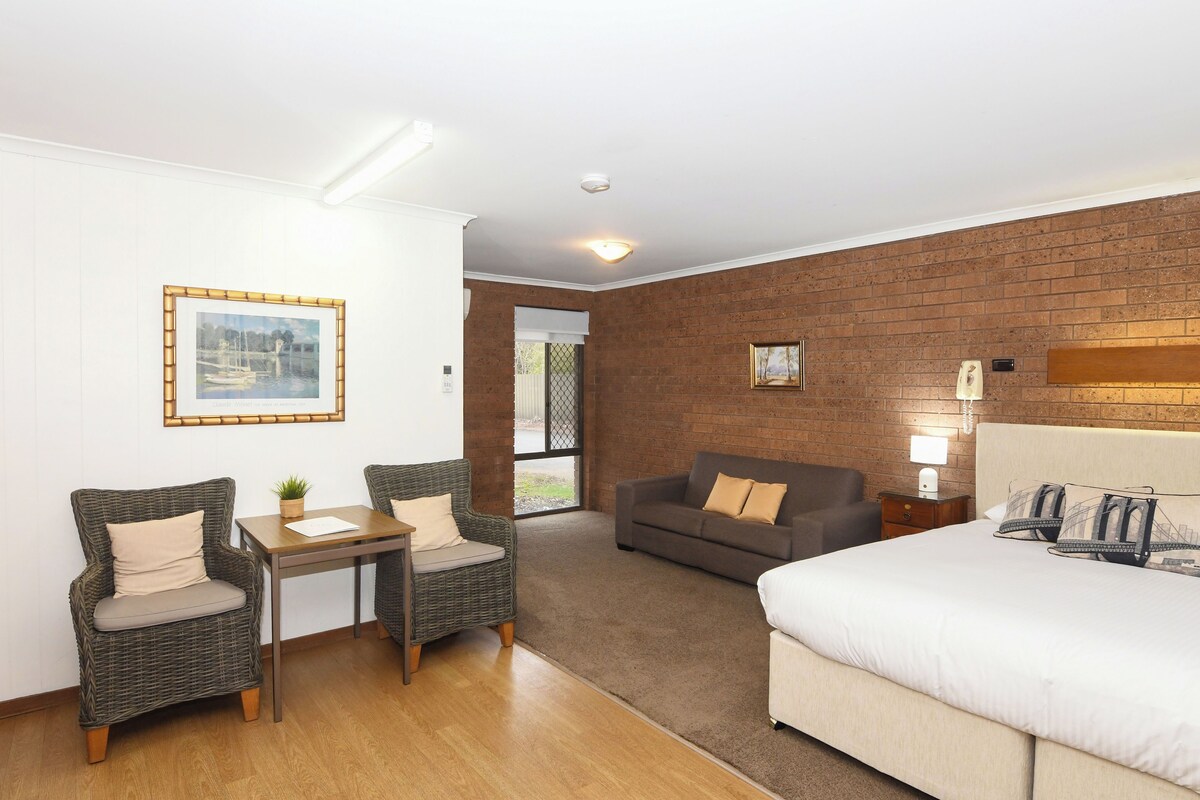 A hotel room with a bed, two wicker chairs, a small table, a sofa, and a brick wall.