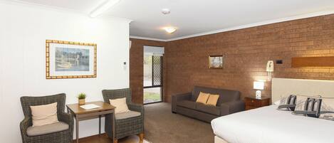 A hotel room with a bed, two wicker chairs, a small table, a sofa, and a brick wall.