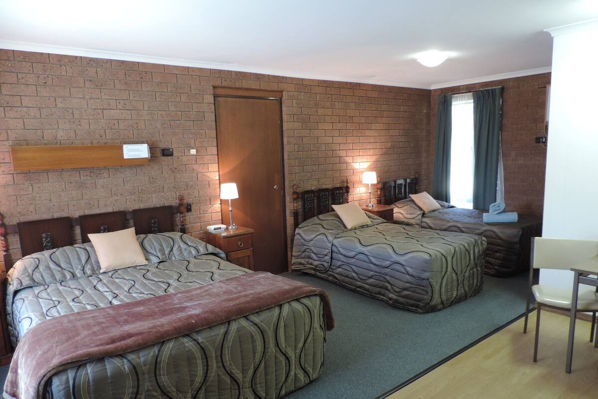 A hotel room with two beds, a wooden headboard, a brick wall, a door, and a small table with a chair.