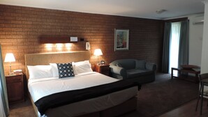 Una habitación de hotel con pared de ladrillo, una cama grande con ropa blanca, dos lámparas de noche, un sillón gris, una mesita y un cuadro en la pared.