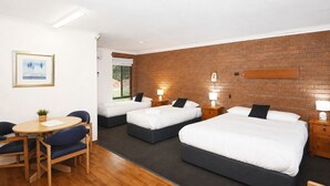 A hotel room with three single beds, a desk with a chair, a small table, and a brick wall. - Greenacres Motel (Corowa)