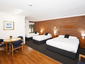 A hotel room with three single beds, a desk with a chair, a small table, and a brick wall. - Greenacres Motel (Corowa)