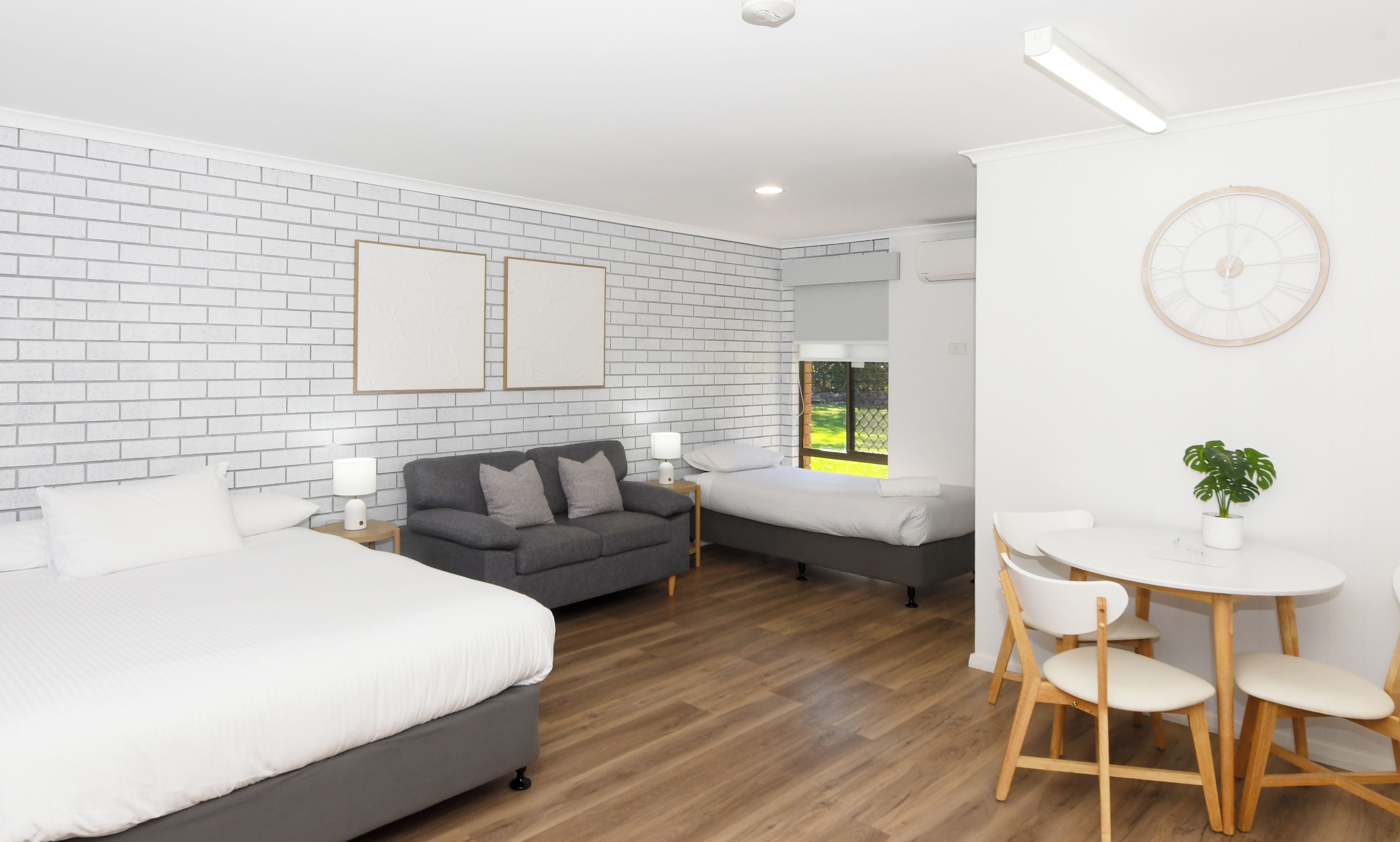 A hotel room with a brick-patterned wall, a large bed, a sofa, a smaller bed, a dining table with chairs, and a potted plant.