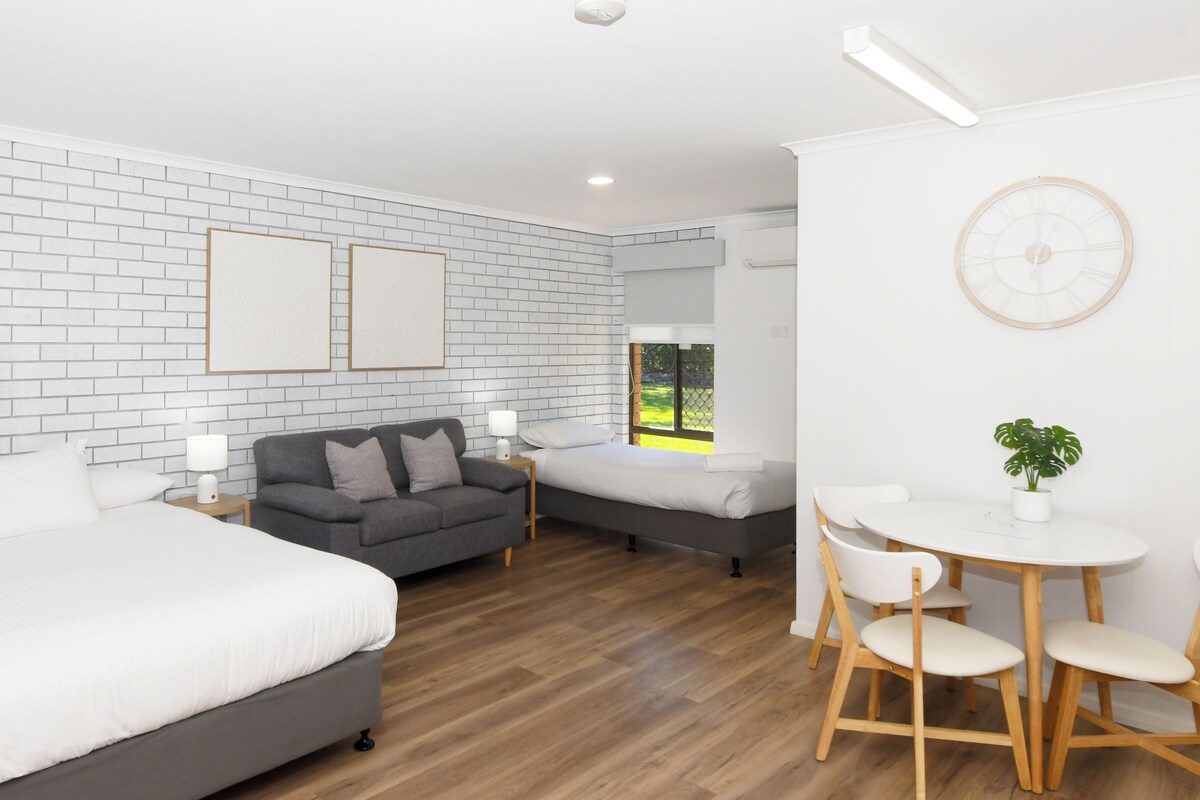 A hotel room with a brick-patterned wall, a large bed, a sofa, a smaller bed, a dining table with chairs, and a potted plant.