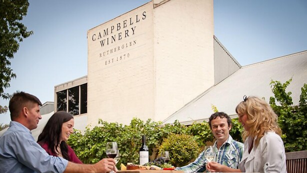 Un grupo de personas sentadas en una mesa al aire libre, con una botella de vino y copas, frente a un edificio que dice 'Campbells Winery'.