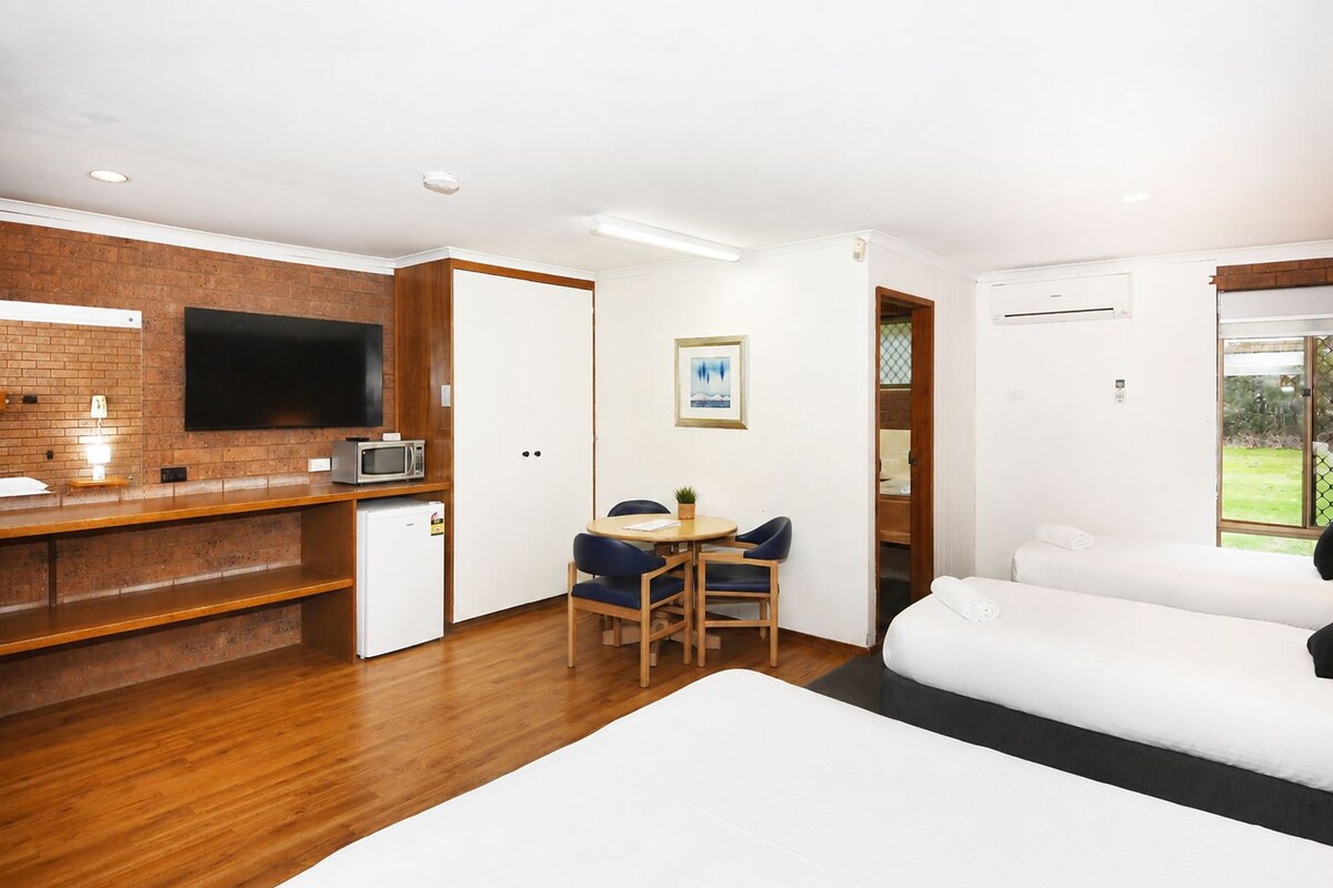 A room with a bed, a desk with a microwave, a chair, and a television mounted on a brick wall.
