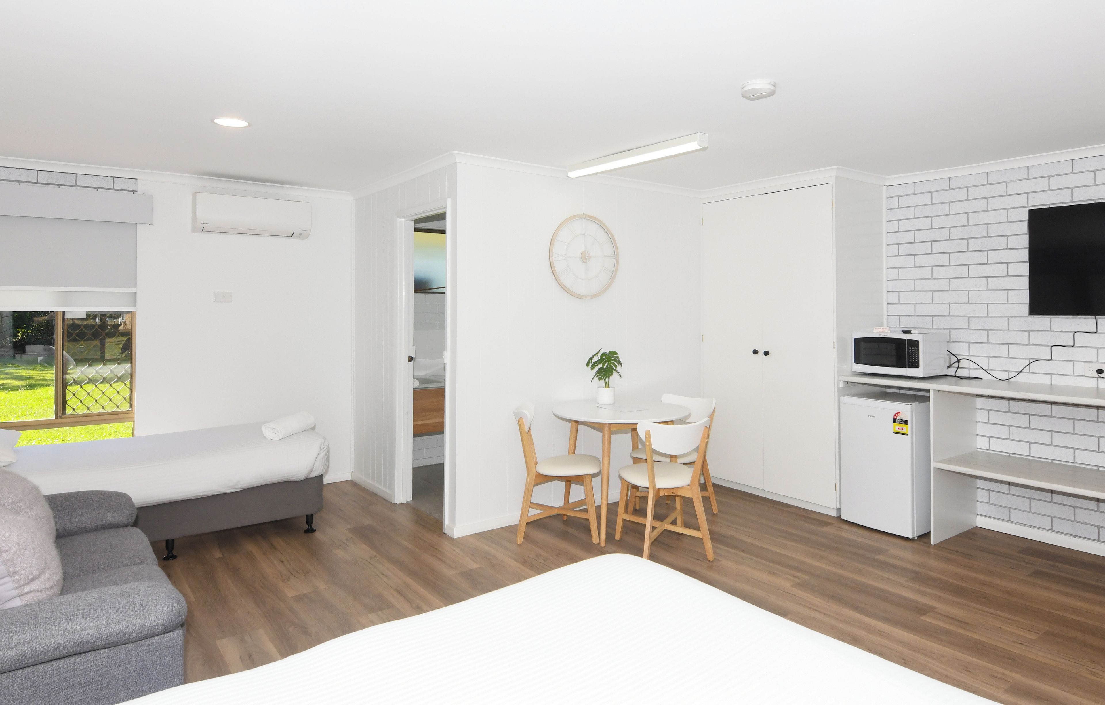 Una habitación moderna y bien iluminada, con una cama, zona de estar con mesa y sillas, una kitchenette pequeña y un televisor montado sobre una pared con diseño de ladrillos.