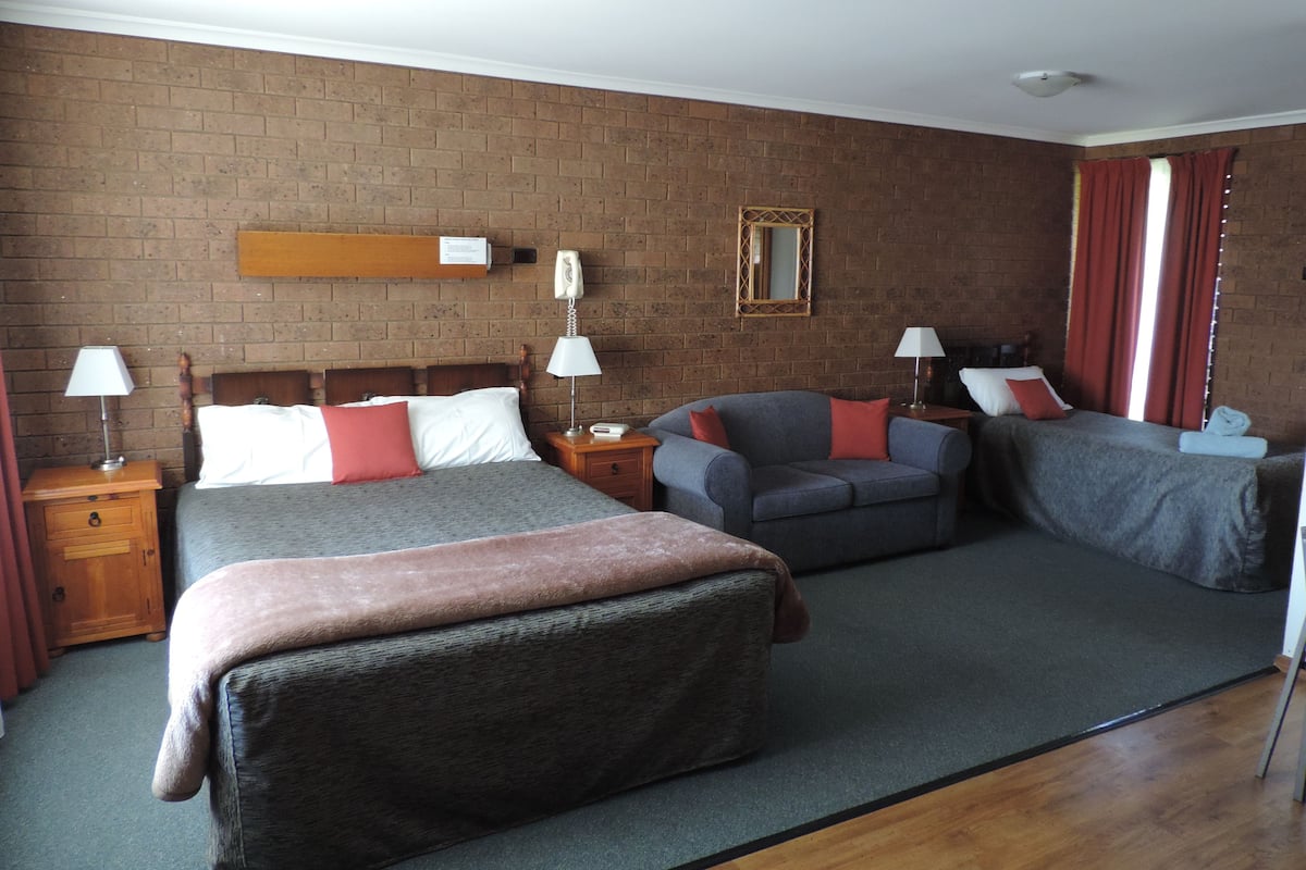 A hotel room with a brick wall, a bed with a wooden headboard, a grey sofa, and a small armchair. There are two bedside tables with lamps and a mirror on the wall.