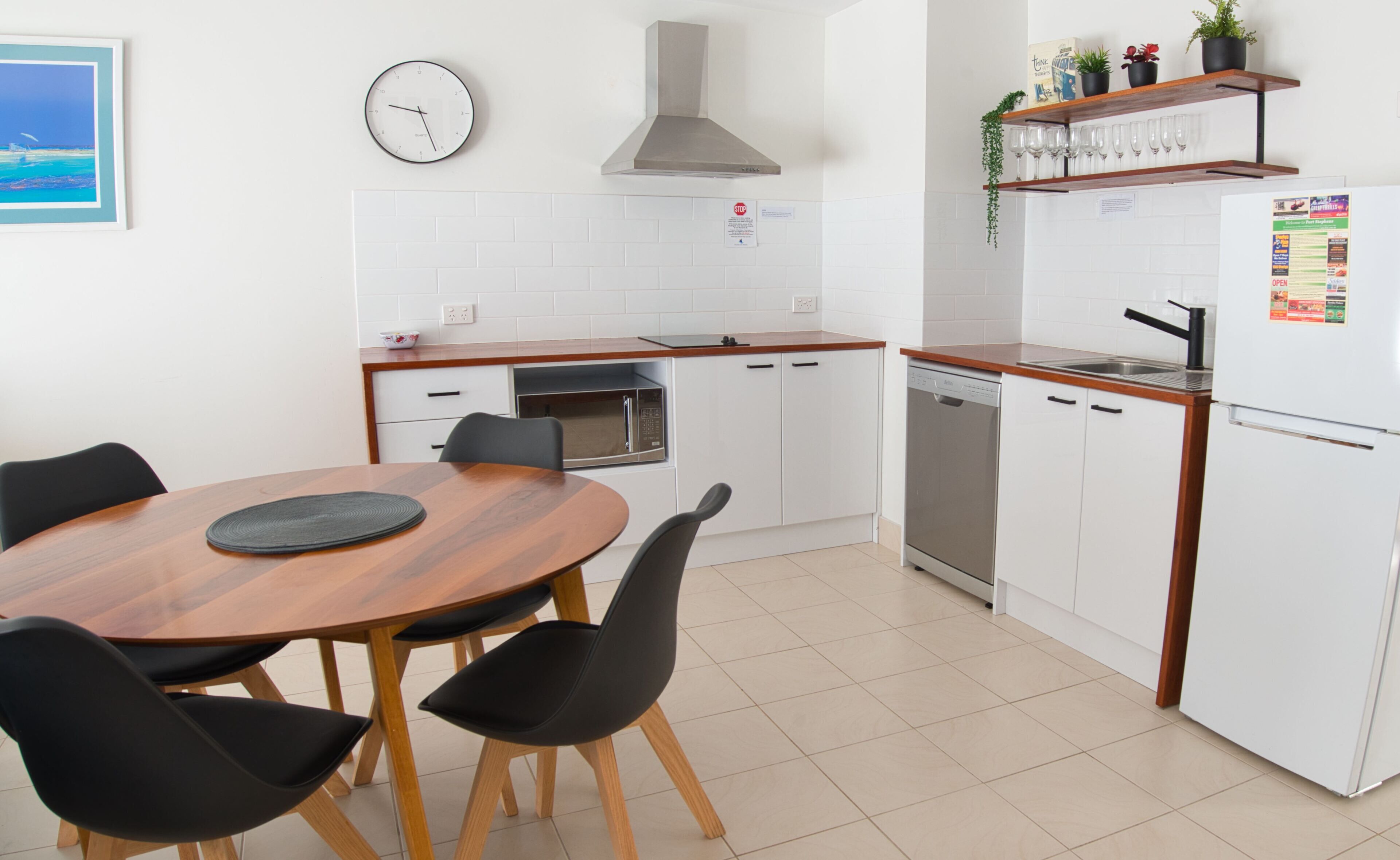A modern kitchen with white cabinets, a wooden dining table, black chairs, a stainless steel range hood, a dishwasher, and a refrigerator with magnets.