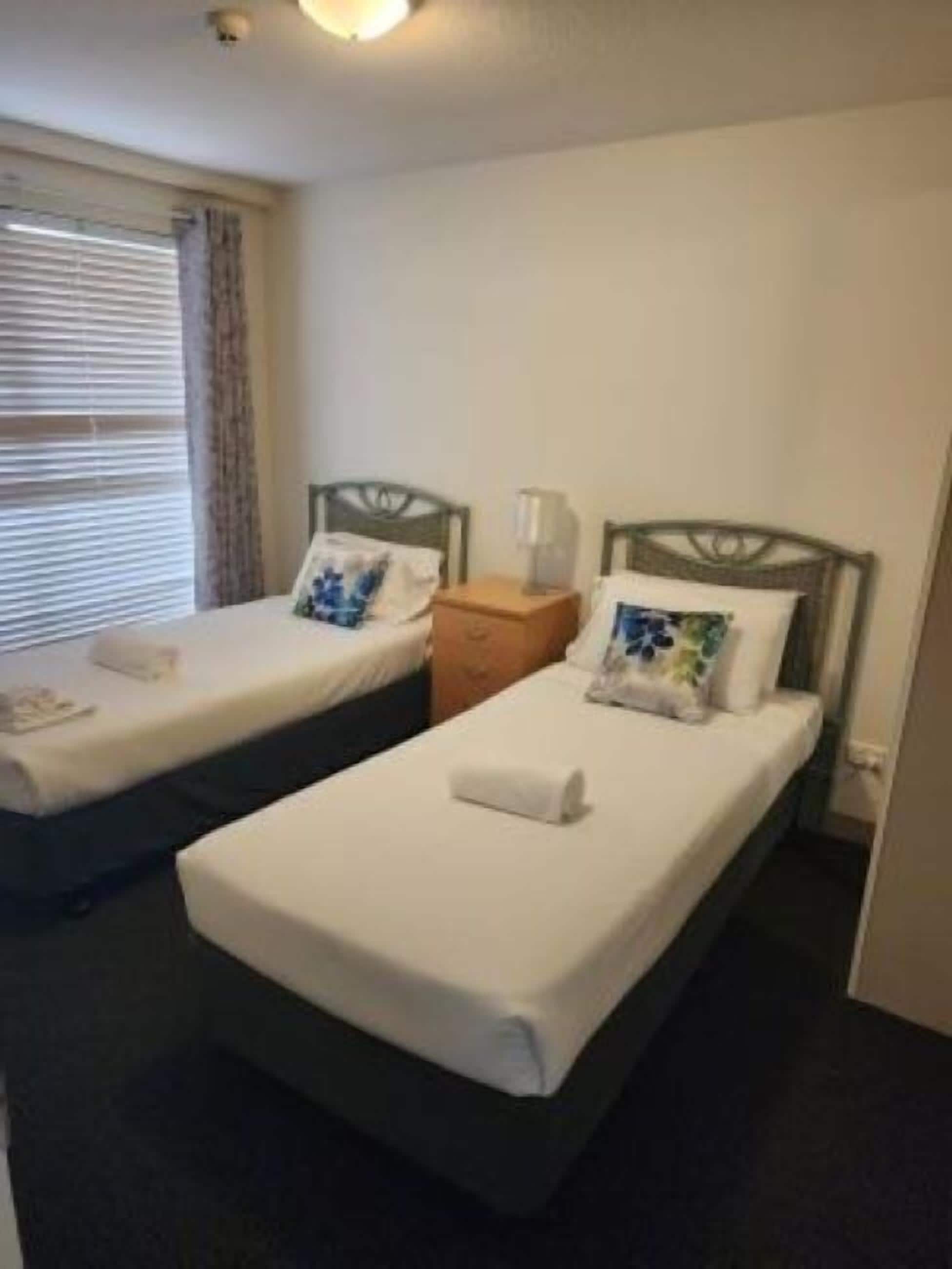 Una habitación de hotel con dos camas individuales, una mesita de noche con lámpara y una ventana con persianas.