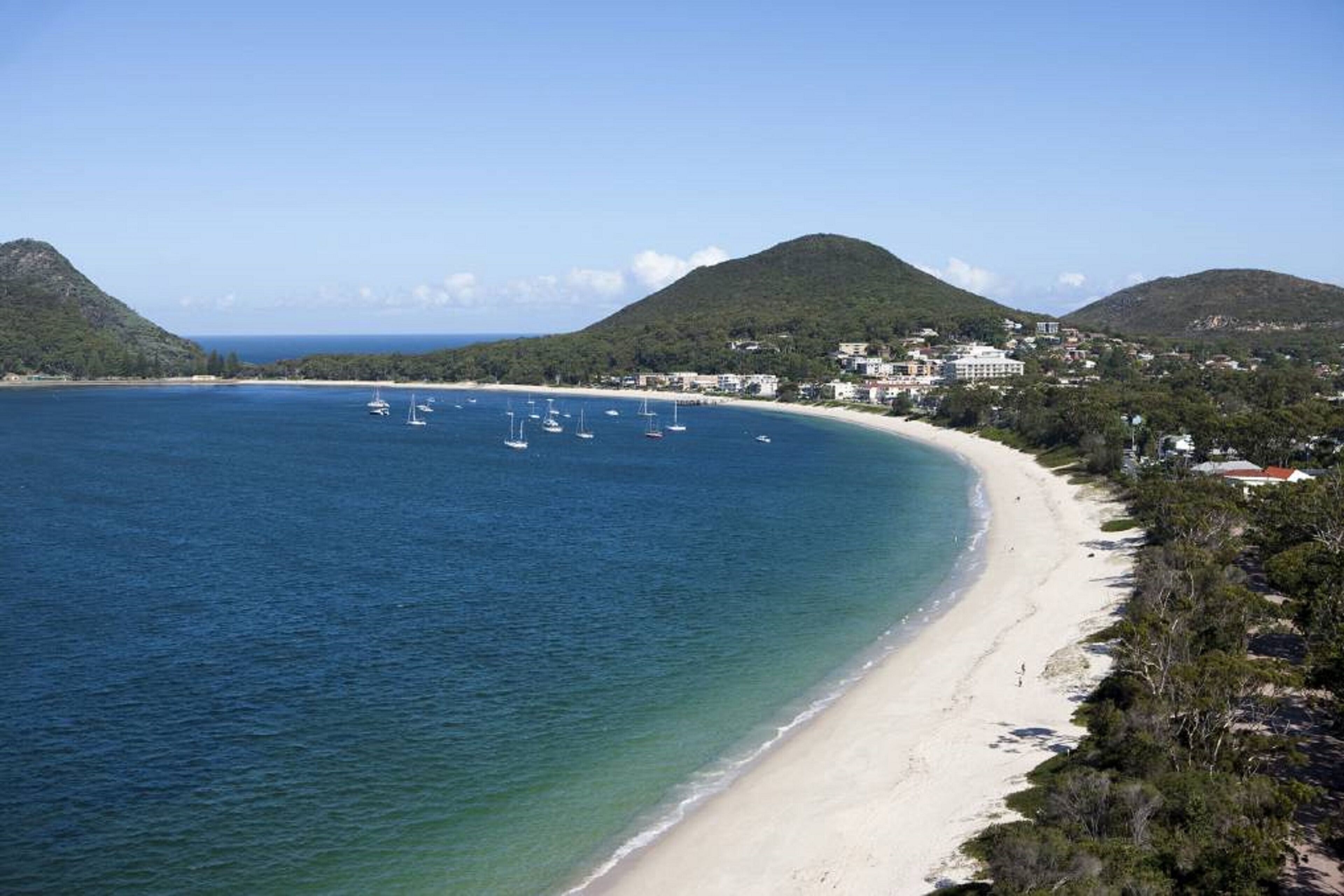 A coastal area with a sandy beach, clear blue waters, and several boats. There are hills in the background and buildings along the shore.