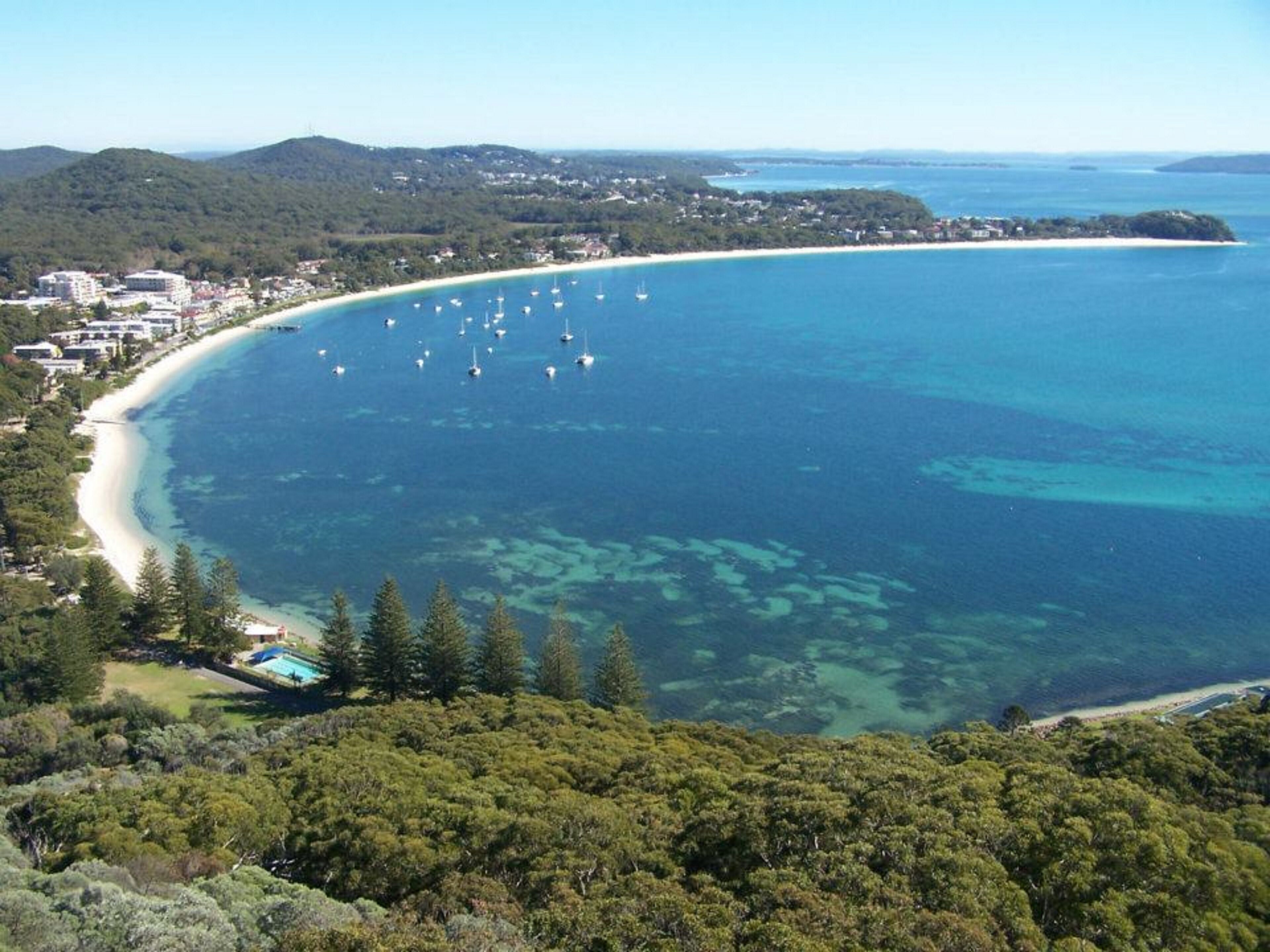 A coastal area with a beach, clear blue waters, and a forested hill.