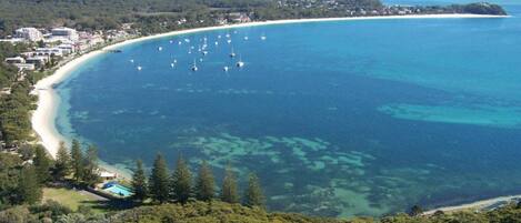 A coastal area with a beach, clear blue waters, and a forested hill.
