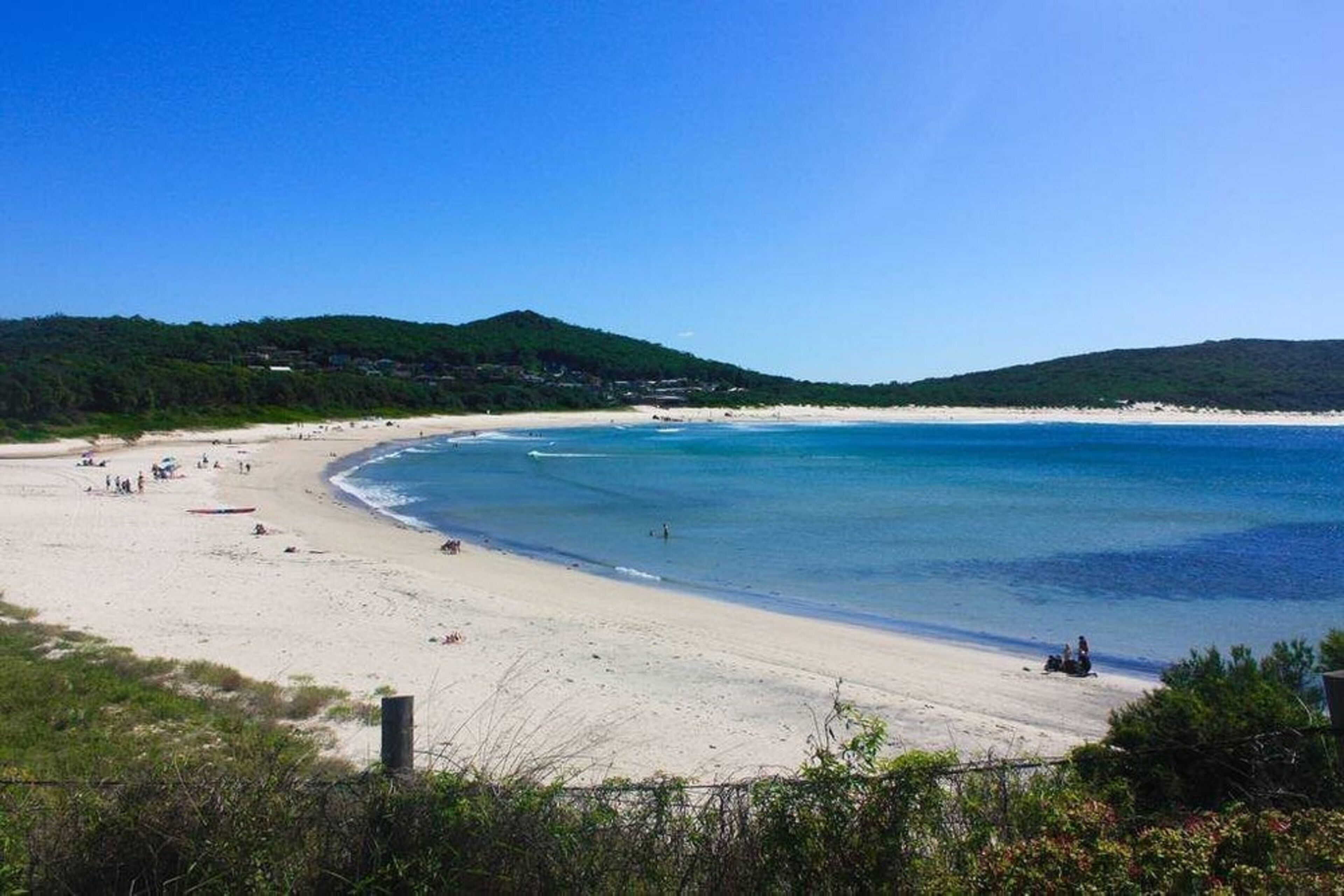 Ein Strand mit klarem, blauem Wasser, sandigem Ufer und grünen Hügeln im Hintergrund.