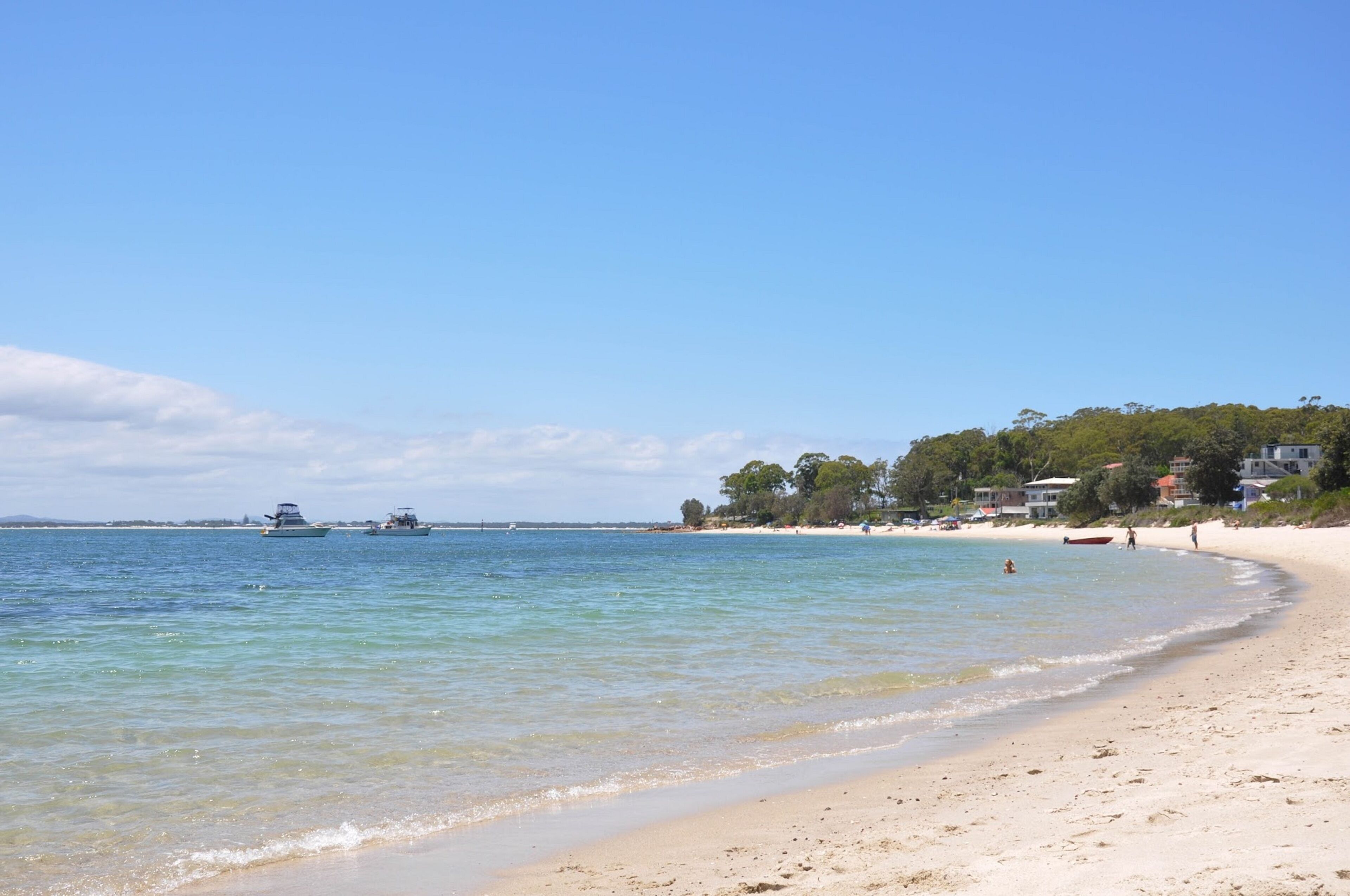 Una playa con agua cristalina, barcos y edificios al fondo.
