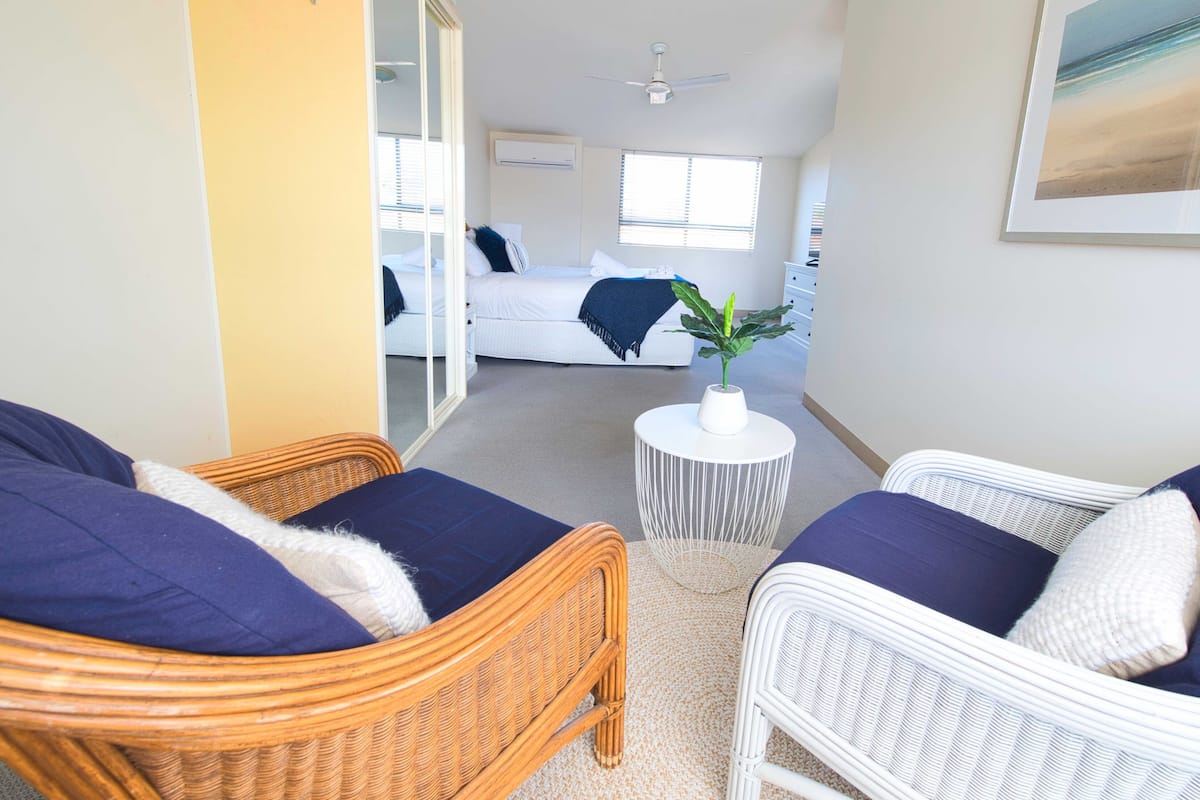 Ein Zimmer mit Korbmöbeln, einem kleinen weißen Tisch mit Pflanze und einem Bett mit blauer Decke.