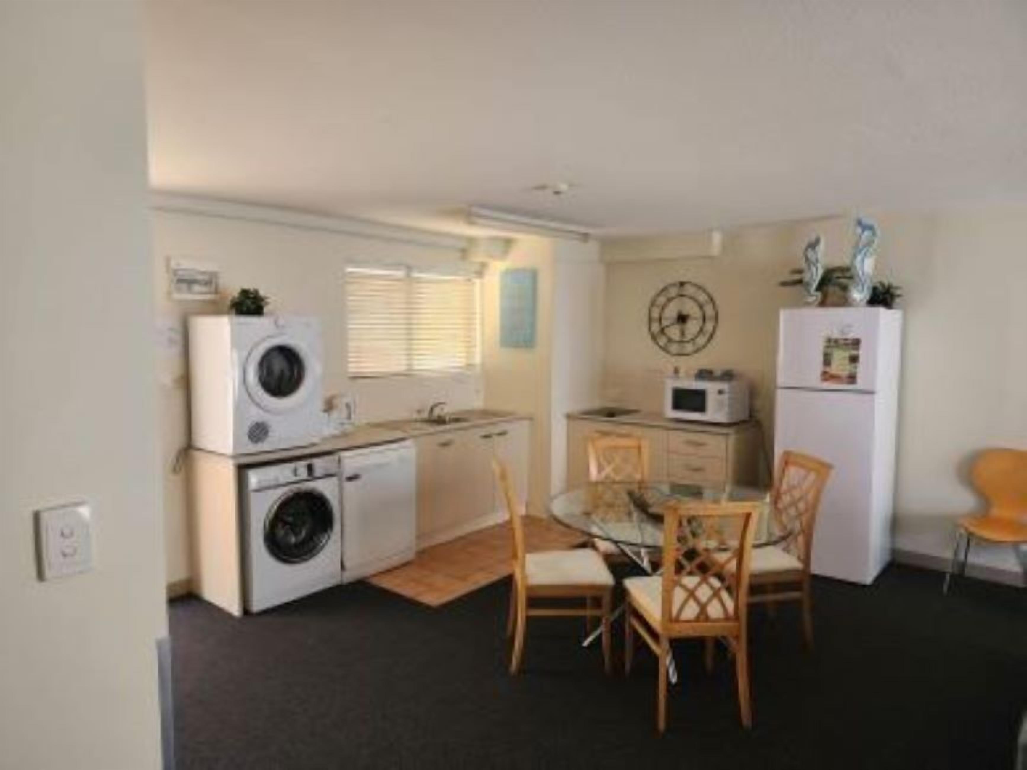 A compact kitchen with a washing machine, microwave, and a small refrigerator. There is a dining table with chairs and a window with blinds.