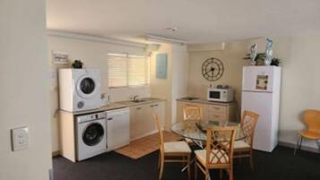 A compact kitchen with a washing machine, microwave, and a small refrigerator. There is a dining table with chairs and a window with blinds.