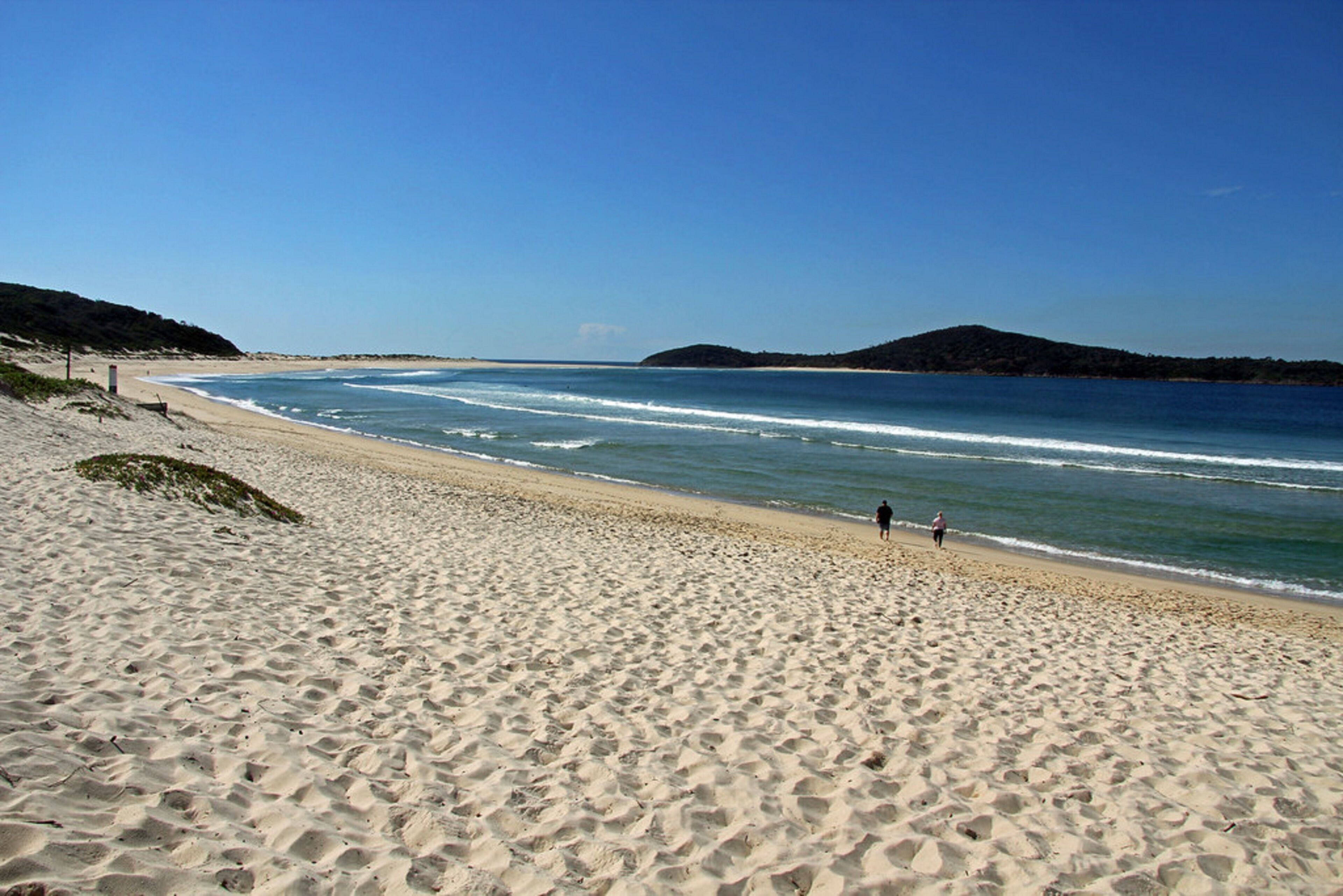 A sandy beach with gentle waves, a clear sky, and distant hills.