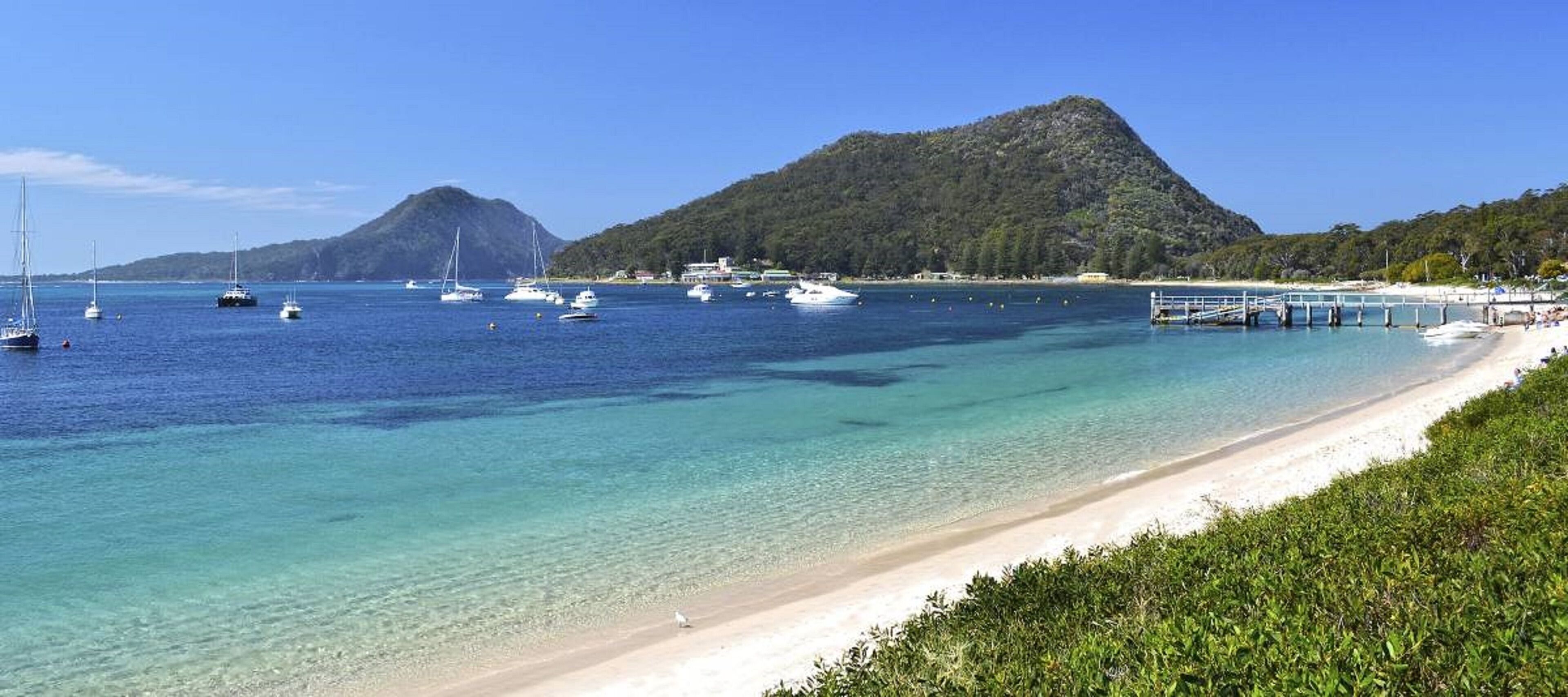 Una spiaggia costiera con acque turchesi cristalline, imbarcazioni e montagne sullo sfondo.