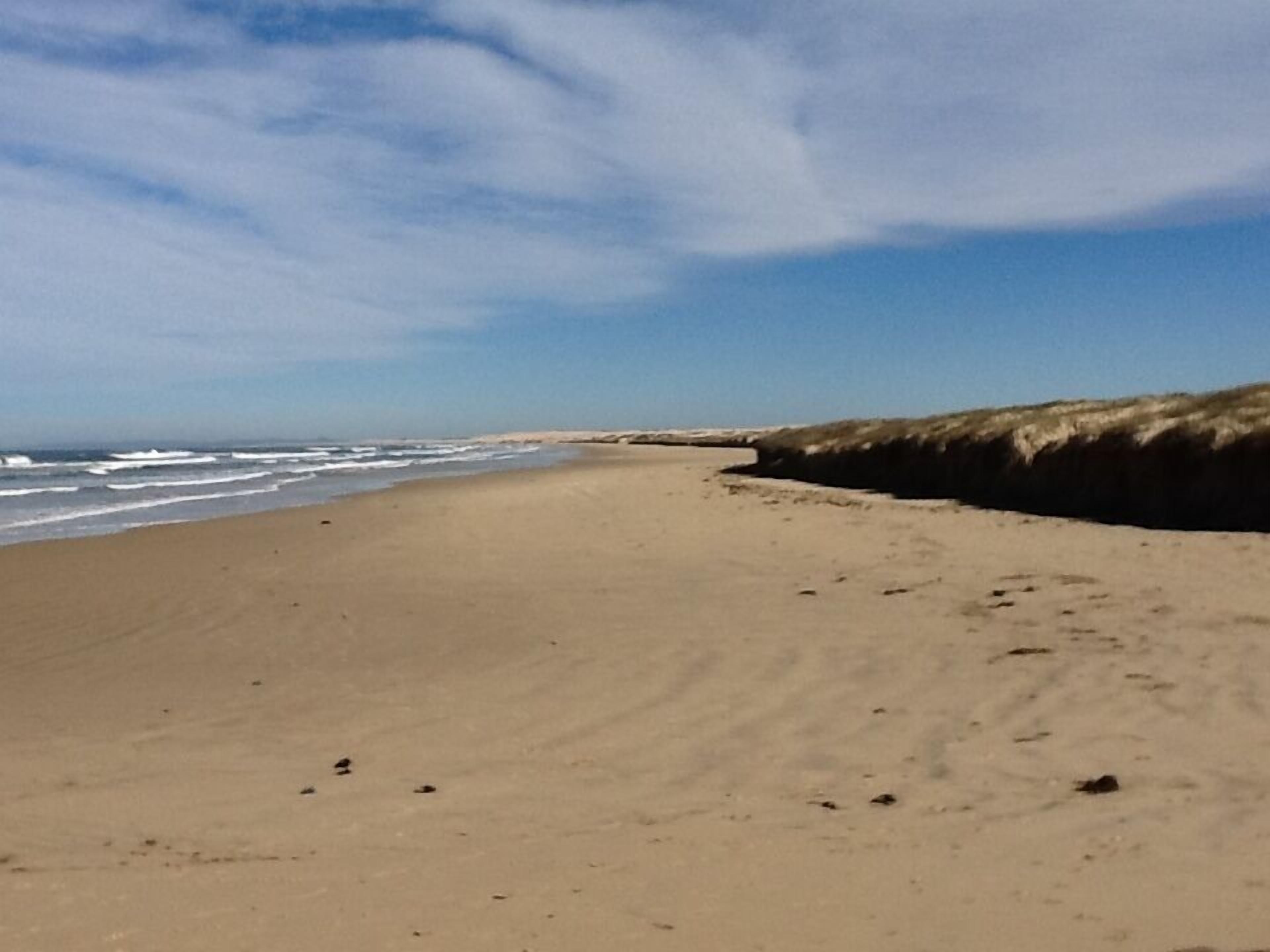 A wide sandy beach with gentle waves and dunes in the background.