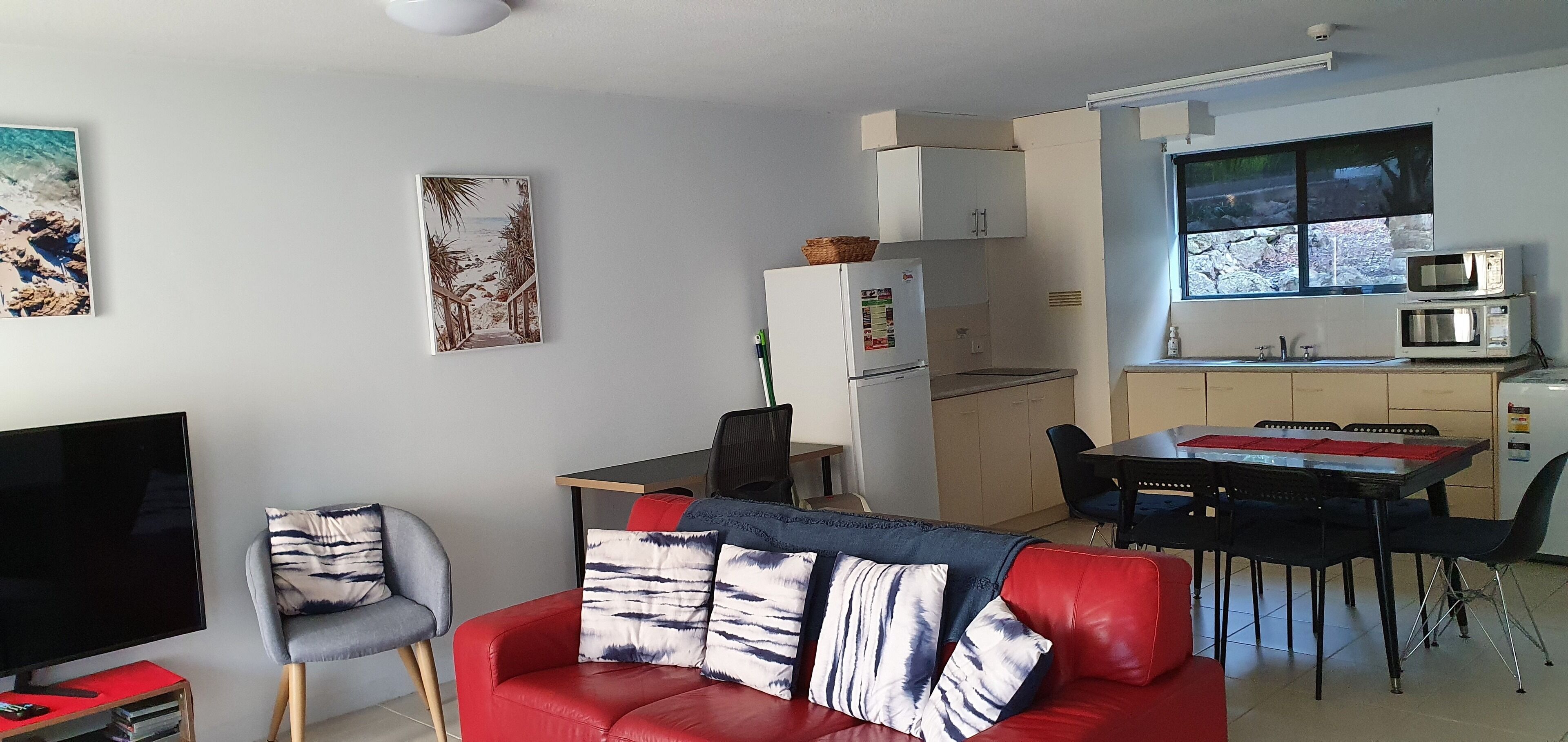 A compact living space with a red sofa, a TV, a dining table with chairs, a kitchen area with a fridge and microwave, and a window with a view.
