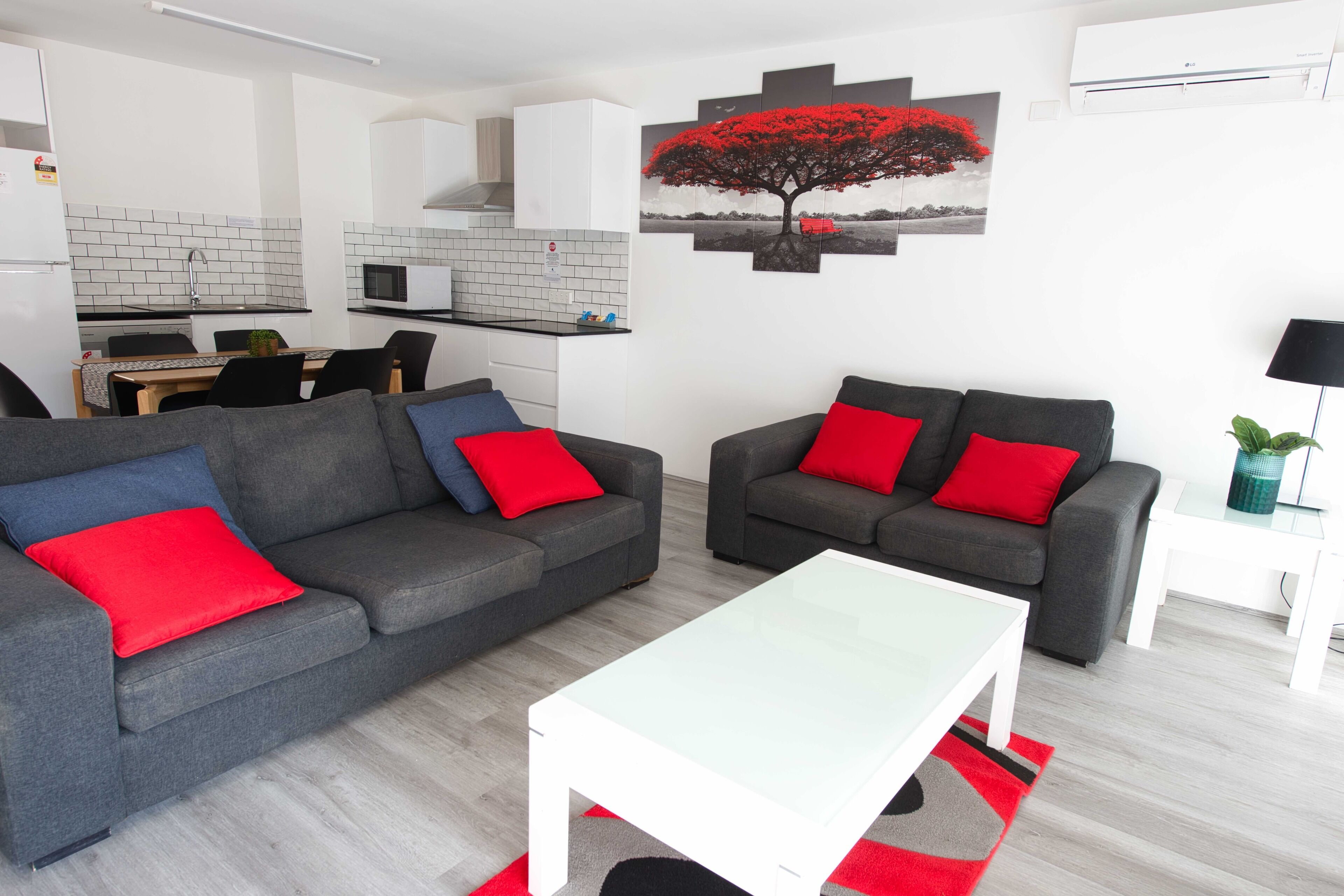 A modern living room with two grey sofas, each with a red cushion, and a white coffee table. A kitchen with white cabinets and a tiled backsplash is visible in the background.