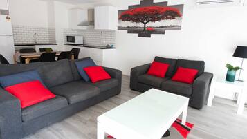 A modern living room with two grey sofas, each with a red cushion, and a white coffee table. A kitchen with white cabinets and a tiled backsplash is visible in the background.