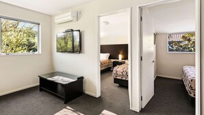 A compact hotel room with a bed, a desk, a chair, a small table, a window, and an air conditioning unit. - Quality Suites Amore (Christchurch)