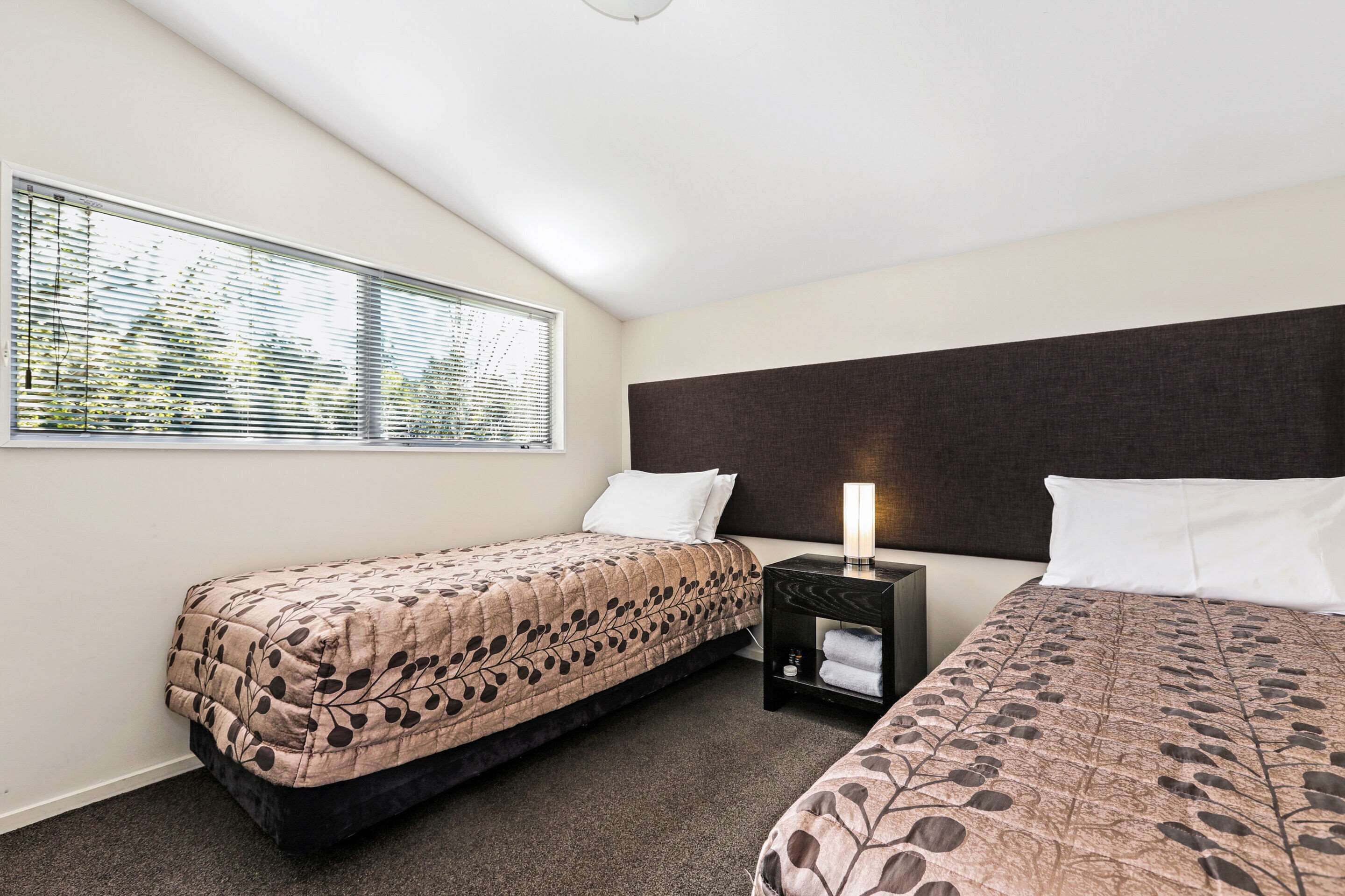 Een hotelkamer met twee bedden, een nachtkastje met een lamp en een raam met jaloezieën.