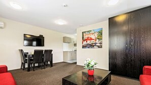 A modern living room with a flat-screen TV, a dining area with chairs and a table, a red sofa, a black coffee table with a plant, and a large dark wooden wardrobe. - Quality Suites Amore (Christchurch)