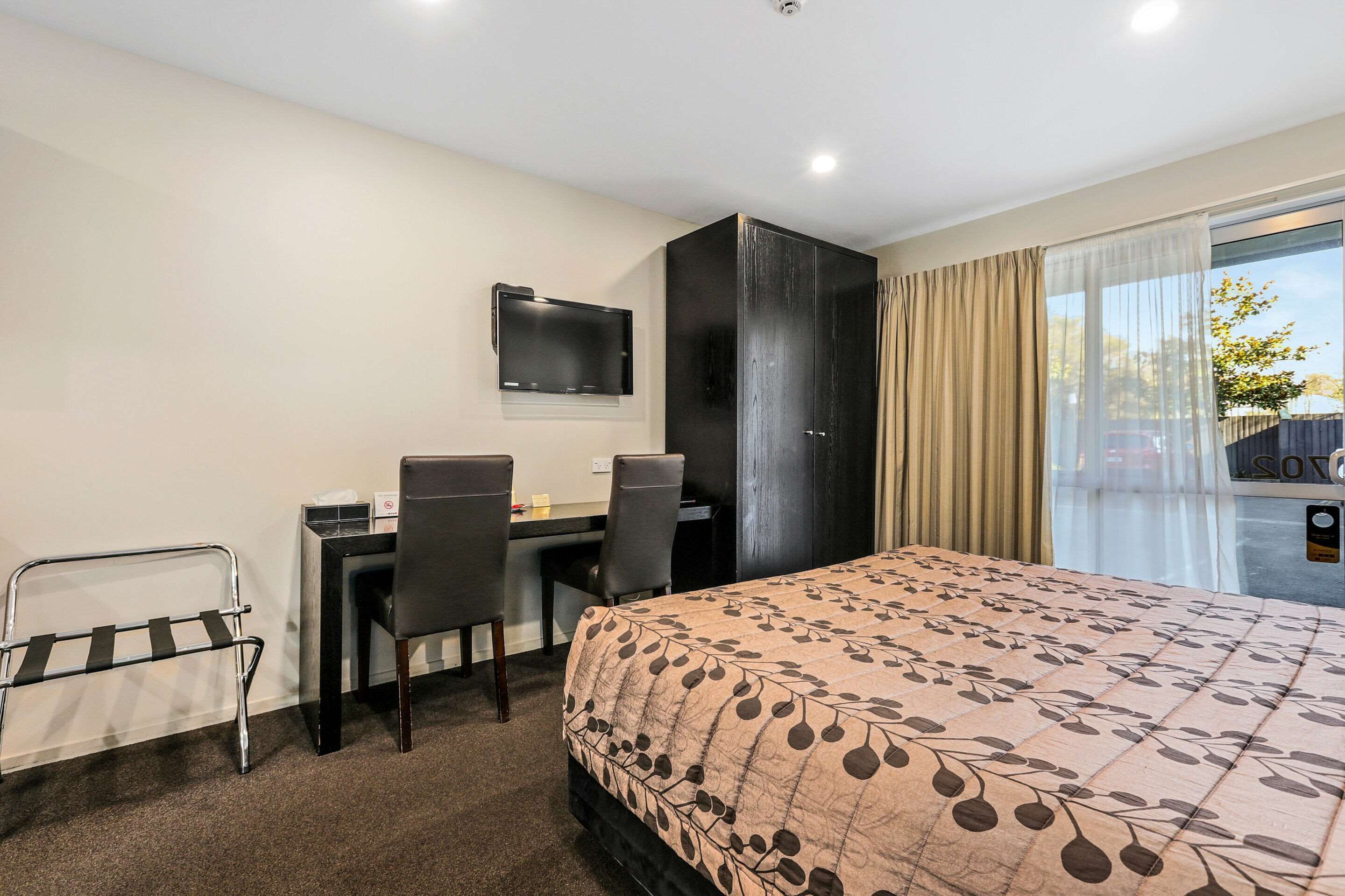 A hotel room with a bed, a desk with two chairs, a TV mounted on the wall, and a sliding glass door to the outside.