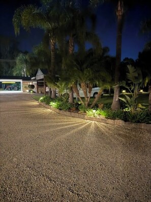 A gravel driveway leading to a building with a garage, surrounded by palm trees and well-lit landscaping. - Barooga River Gums Motor Inn (Barooga)