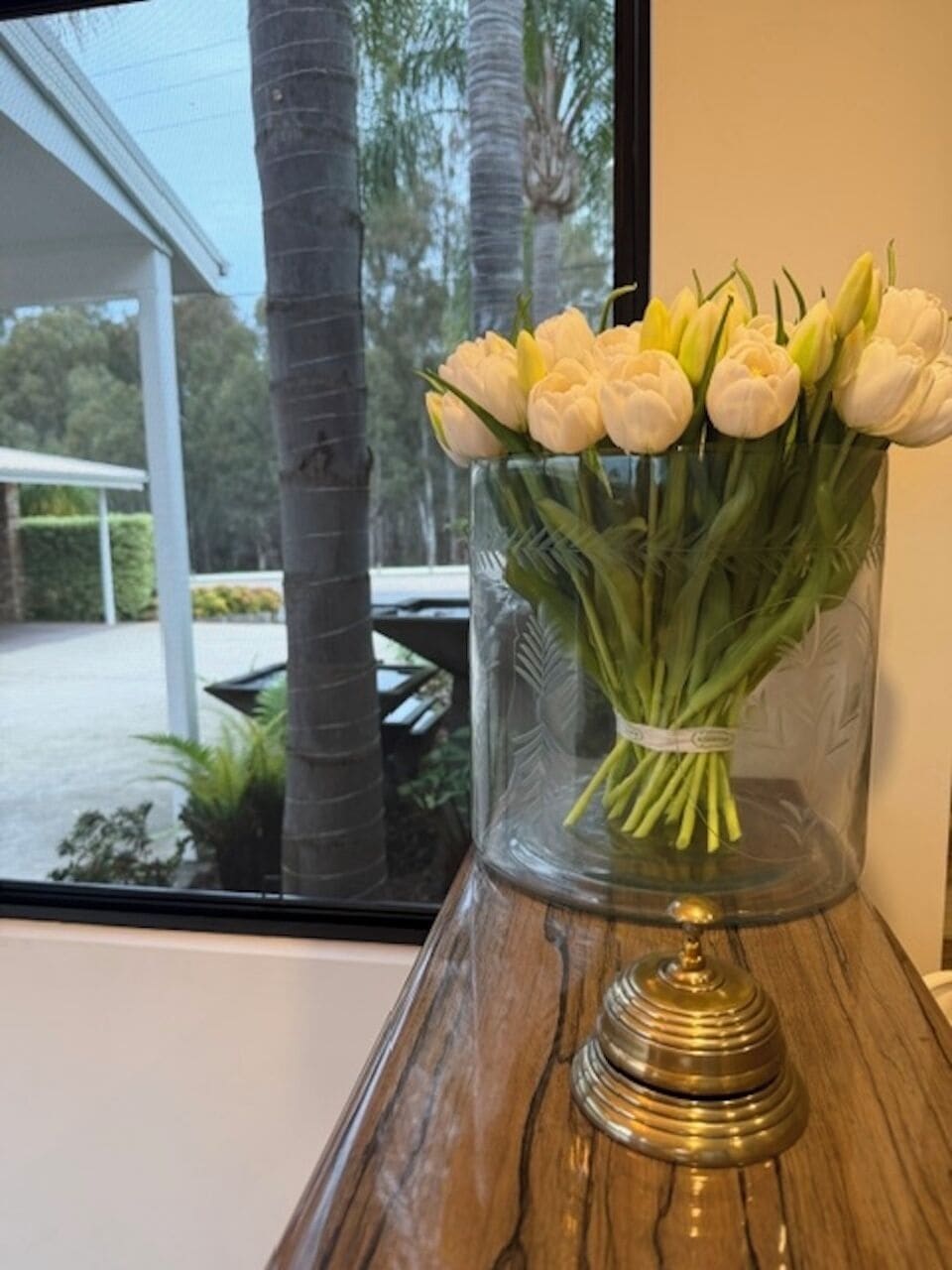 Eine Vase mit weißen Blumen auf einer Holztischplatte nahe eines Fensters.