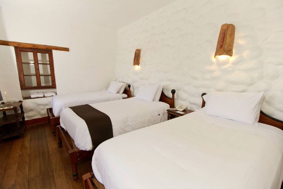 A hotel room with three single beds, wooden headboards, a wooden table, and a window with wooden frames.
