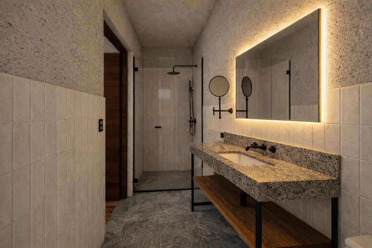 A modern bathroom with a granite countertop sink, a wooden bench, a walk-in shower, and a large mirror with wall-mounted lights.
