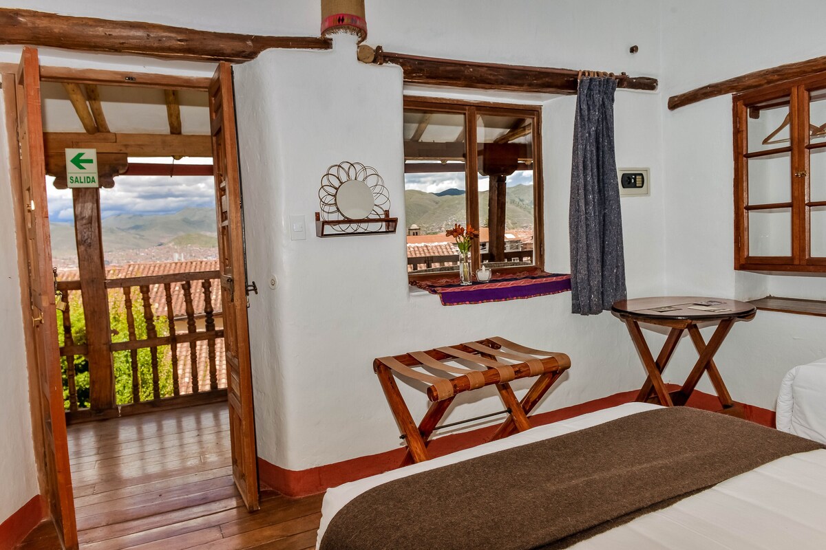A room with a bed, a small table, a bench, a window with a view of mountains, and a door with a sign.