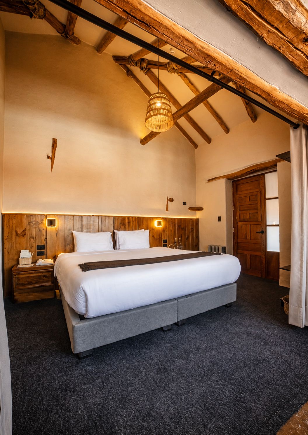 A bedroom with a large bed, wooden beams, a hanging light fixture, and a wooden door.