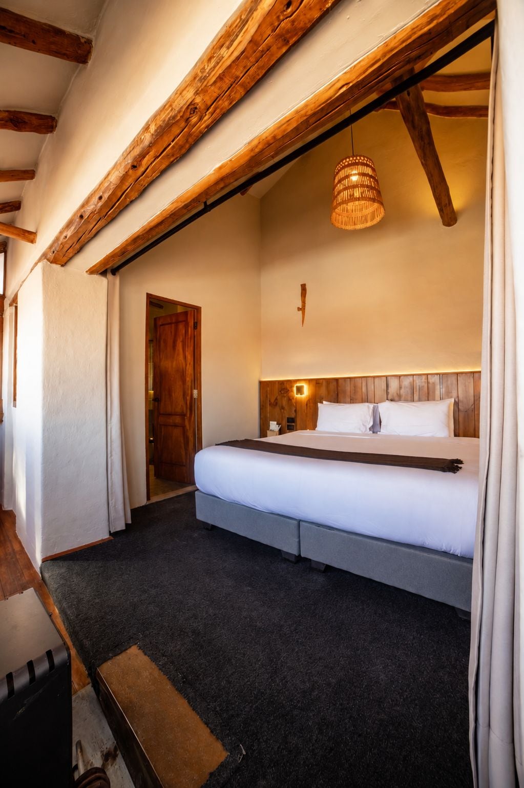 A bedroom with a bed, wooden beams, a hanging light fixture, and a door.