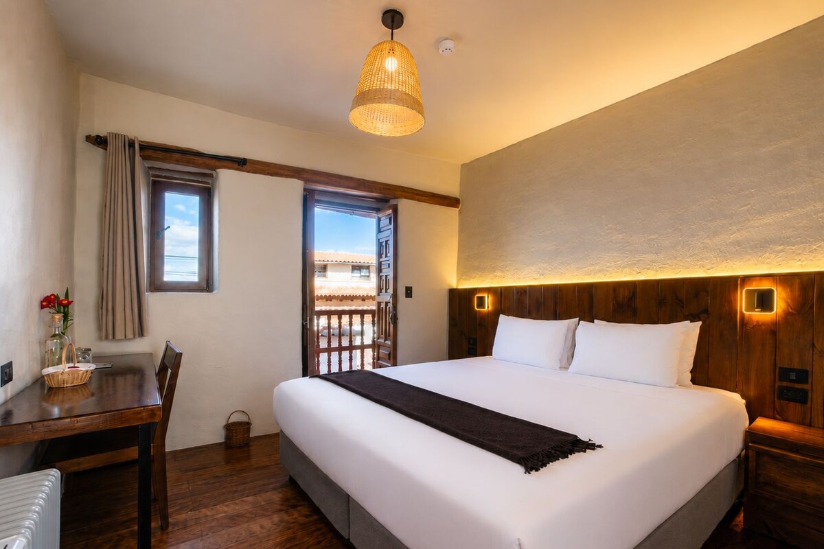 A hotel room with a large bed, a wooden headboard, a desk with a chair, a window with curtains, and a balcony.