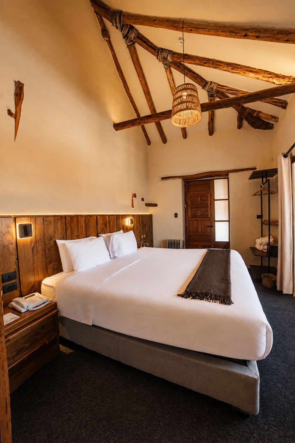 Una habitación de hotel con una cama grande, vigas de madera, una lámpara colgante, un armario de madera y una puerta que da a otra habitación.