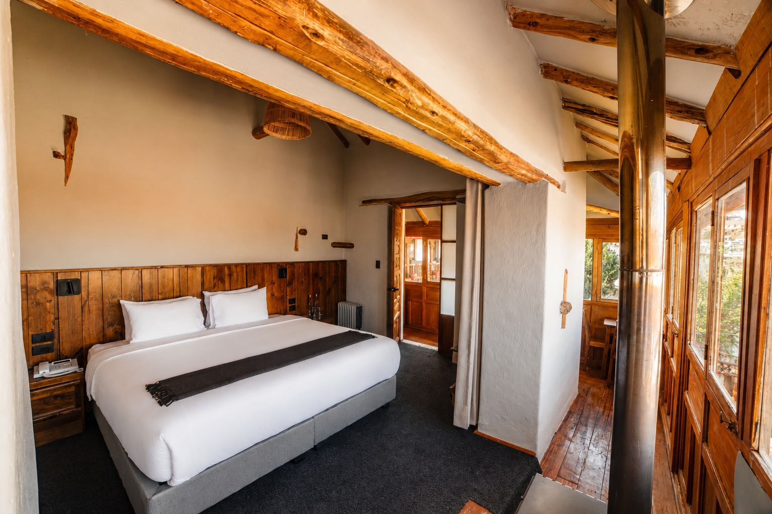 Habitación de hotel con cabecera de madera, cama grande, mesita de noche y vista por la ventana.