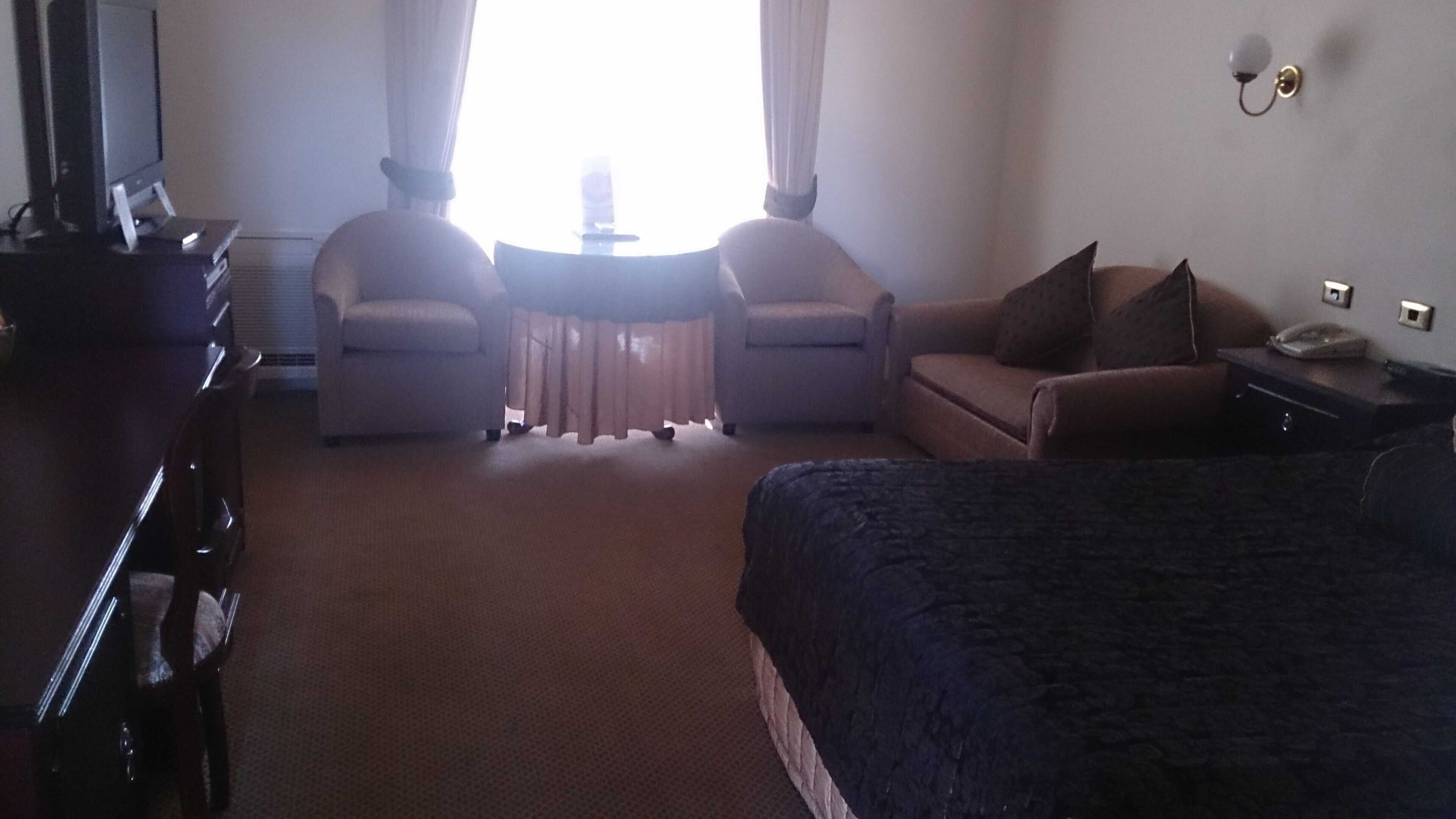 A hotel room with a bed, two armchairs, a small table, a desk, and a television.