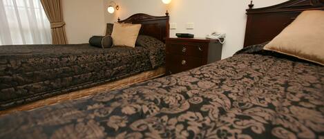 A hotel room with two beds, each with a patterned bedspread, a wooden nightstand with a lamp, and a window with curtains.