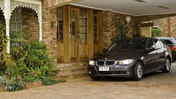 A black BMW parked in a driveway with a brick house, wooden doors, and a white porch railing. - Australian Heritage Motor Inn (Dubbo)