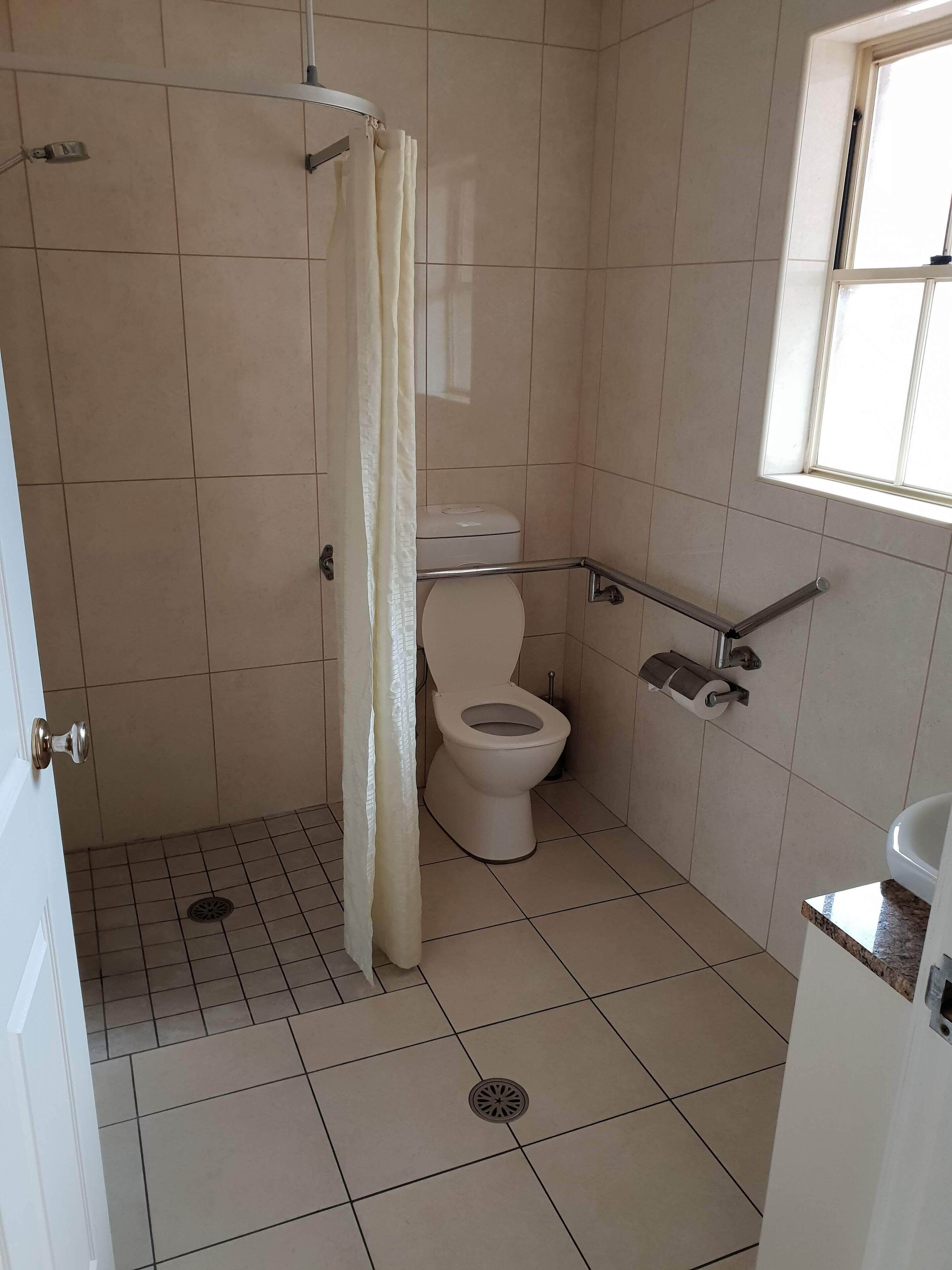 A small, tiled bathroom with a toilet, a shower curtain, a handrail, and a window.