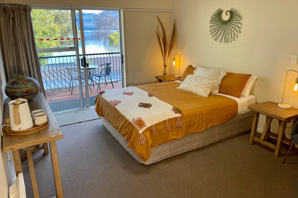 A hotel room with a bed, a desk with a vase, a lamp, and a view of a balcony with a table and chairs.