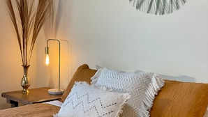 A neatly made bed with a brown blanket, two pillows, a decorative wall hanging, a tall vase with dried plants, and a floor lamp with a lit bulb. - Lakewood Lofts (Te Anau)