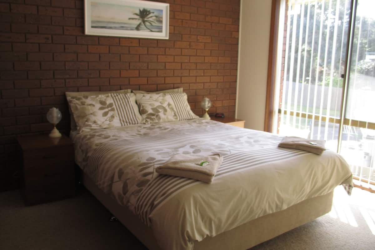 A bedroom with a brick wall, a bed with floral bedding, a nightstand with a lamp, and a window with blinds.